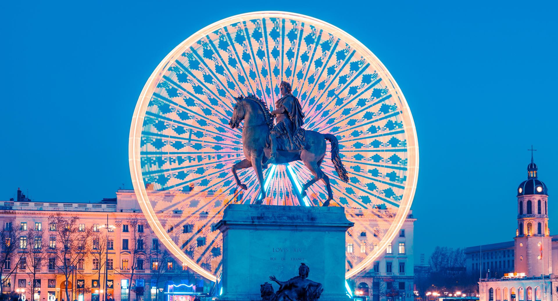 Place de la ville illuminée au crépuscule, avec une grande roue illuminée derrière une statue équestre.