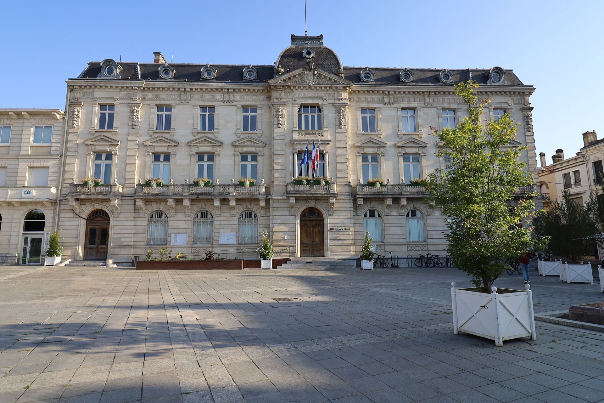 Grand édifice civique en pierre, orné de drapeaux, sur une place ensoleillée, avec un arbre au premier plan.