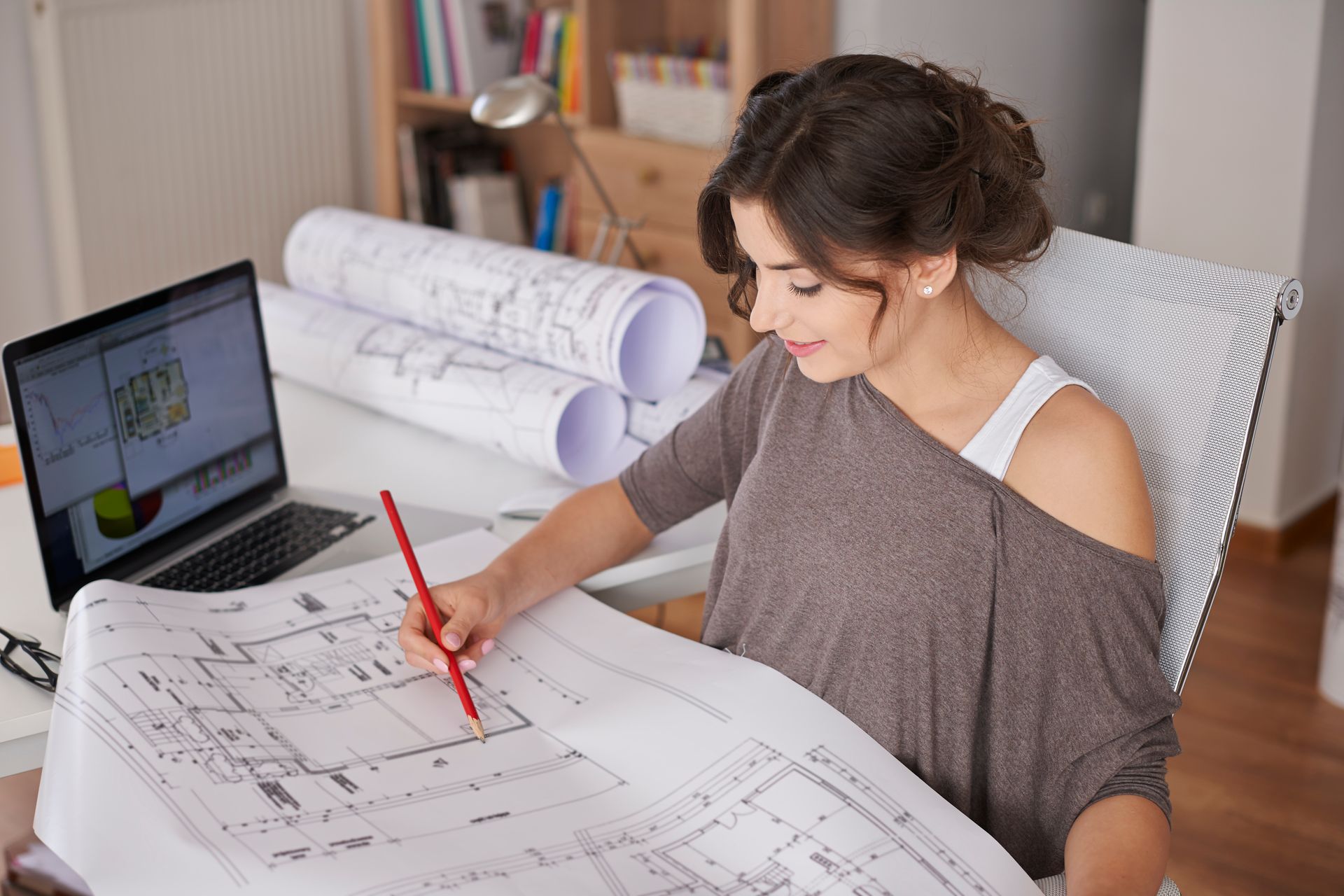 Une femme utilise un crayon rouge pour examiner des plans architecturaux à côté d'un ordinateur portable dans un bureau à domicile.