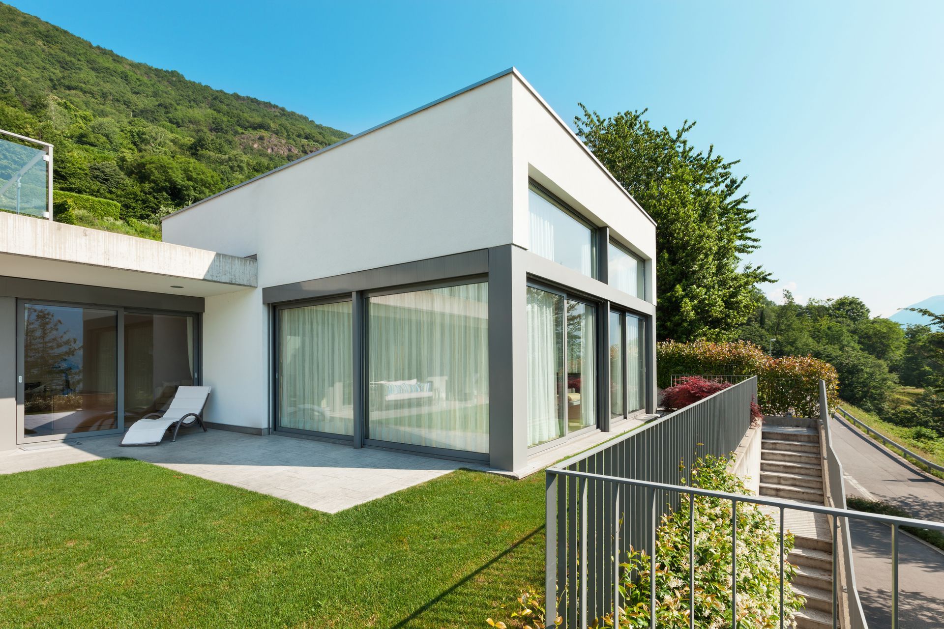 Maison blanche moderne avec de grandes fenêtres, pelouse verdoyante et vue sur la colline, attenante à une terrasse et une clôture.