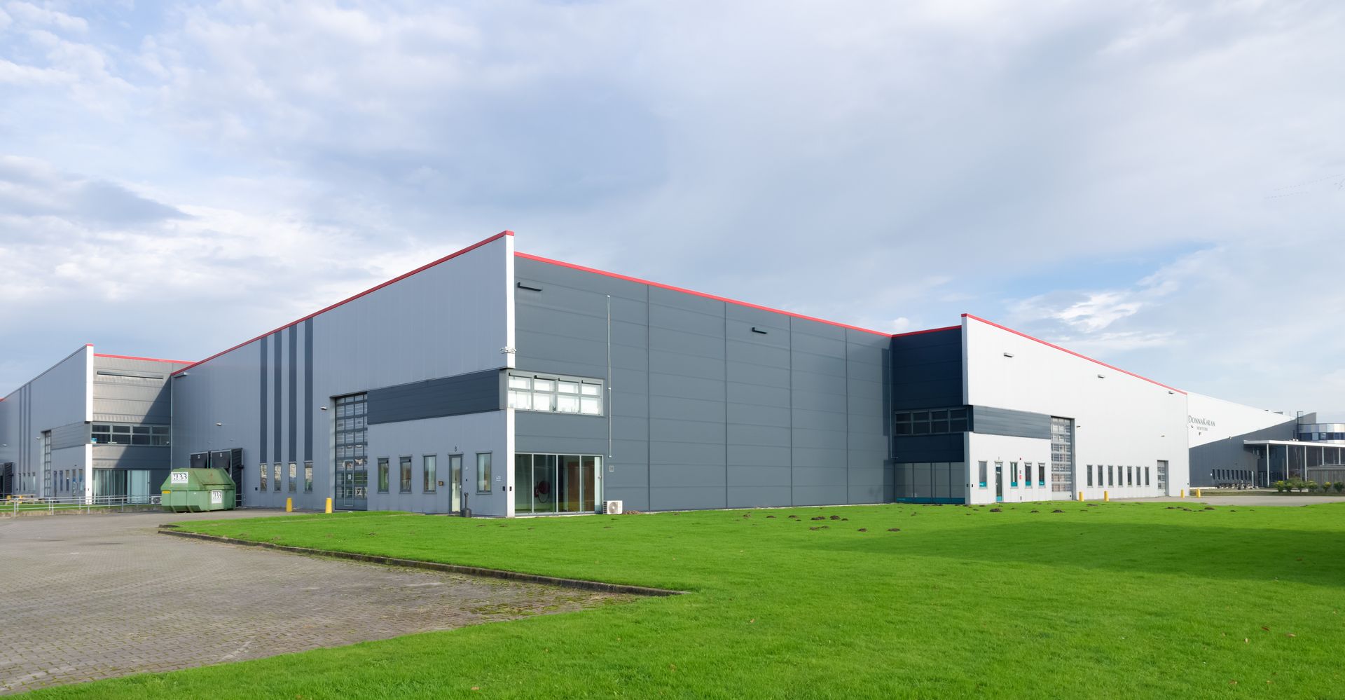 Entrepôt industriel moderne aux panneaux gris et au toit rouge, situé à côté d'une pelouse verte sous un ciel nuageux.