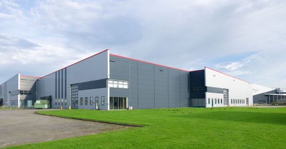 Entrepôt industriel moderne aux panneaux gris et au toit rouge, situé à côté d'une pelouse verte sous un ciel nuageux.