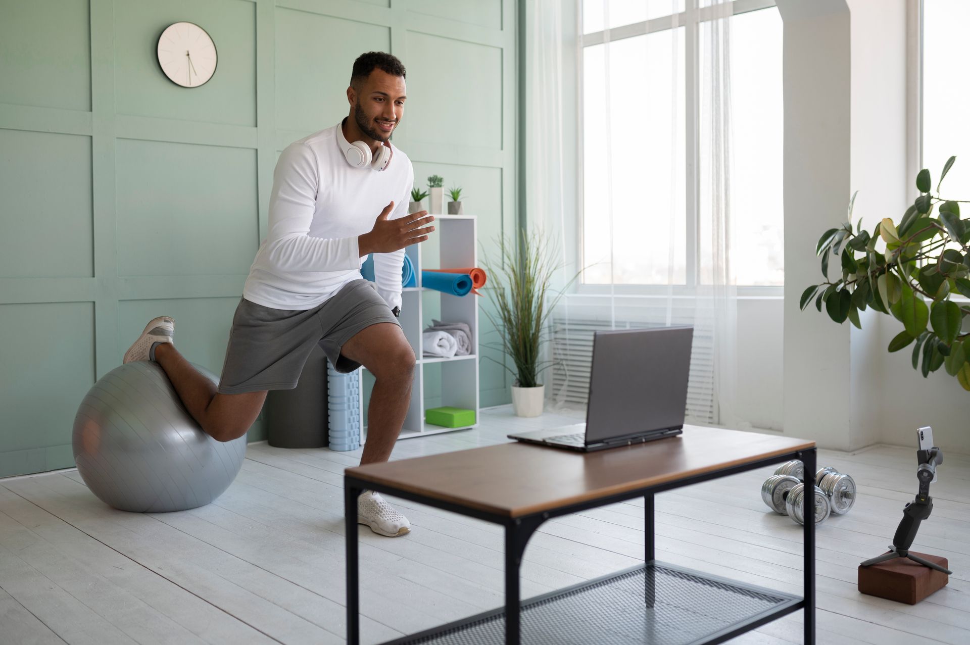 Hombre haciendo una estocada con una pelota de ejercicios, mirando una computadora portátil