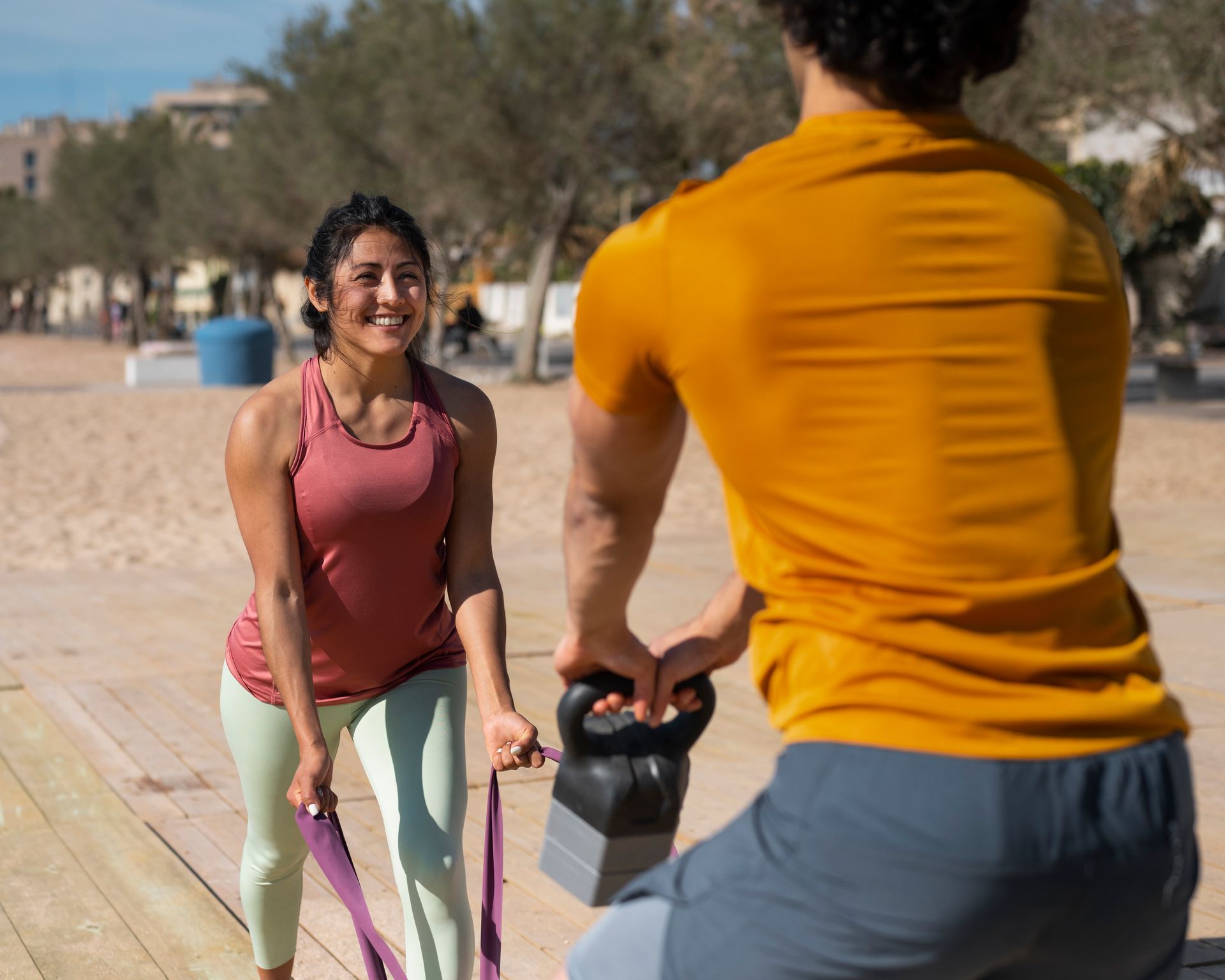 Mujer usando una banda de resistencia, sonriendo a un hombre que sostiene una pesa rusa, en un entorno de playa.