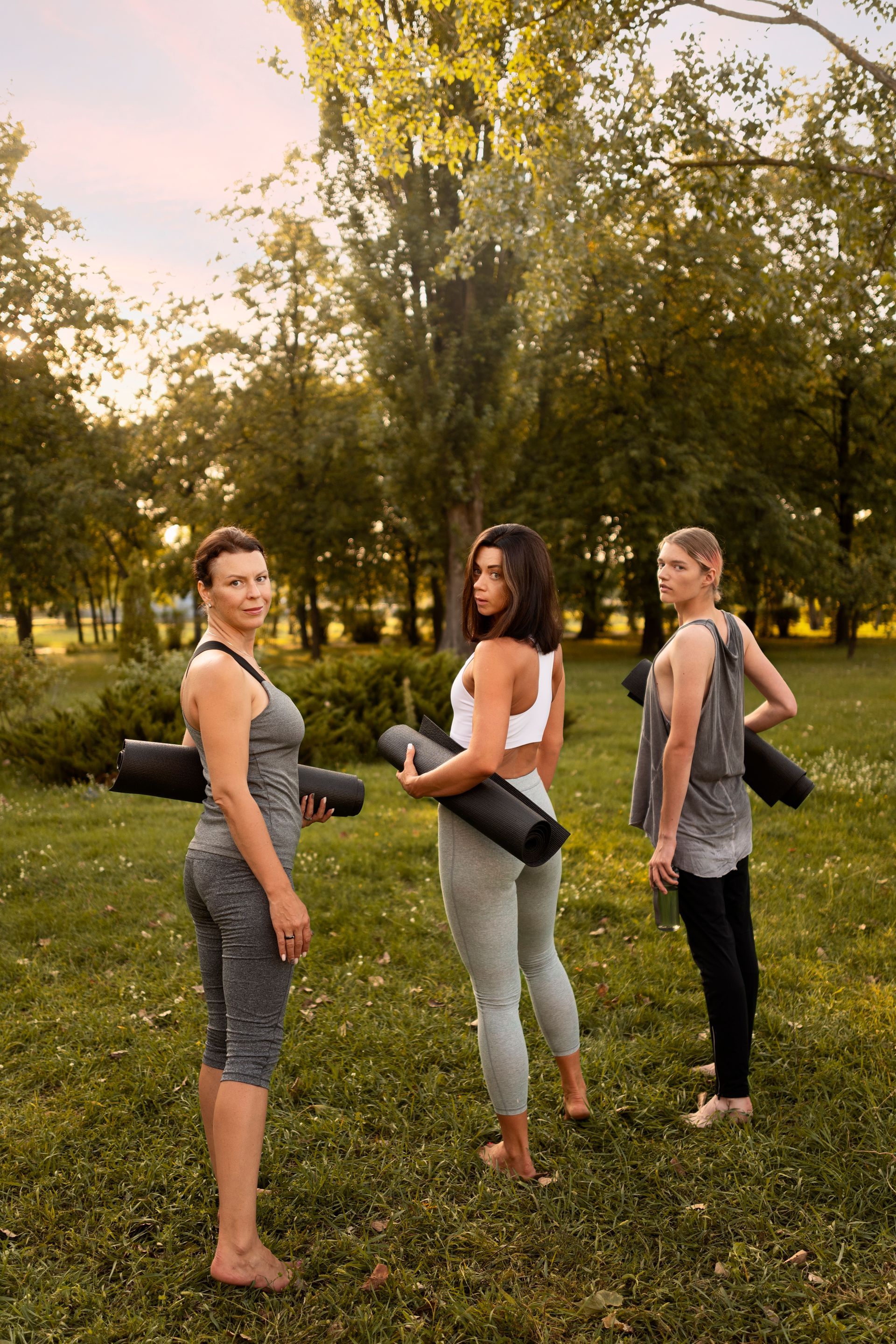 Tres mujeres en ropa deportiva, sosteniendo colchonetas de yoga, en un parque con césped.