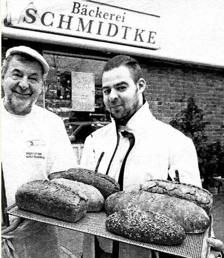 Zwei Männer halten ein Tablett mit Brot vor einem Schild mit der Aufschrift „Bäckerei Schmidtke“