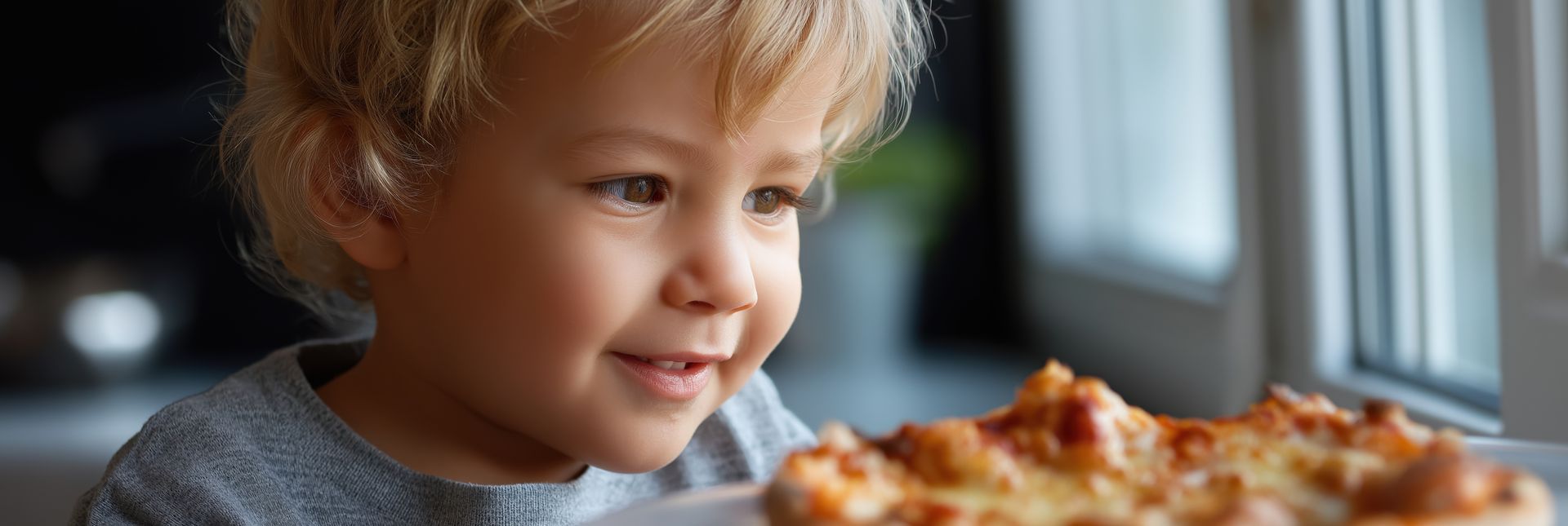 Un enfant souriant regarde une part de pizza, assit près d'une fenêtre.