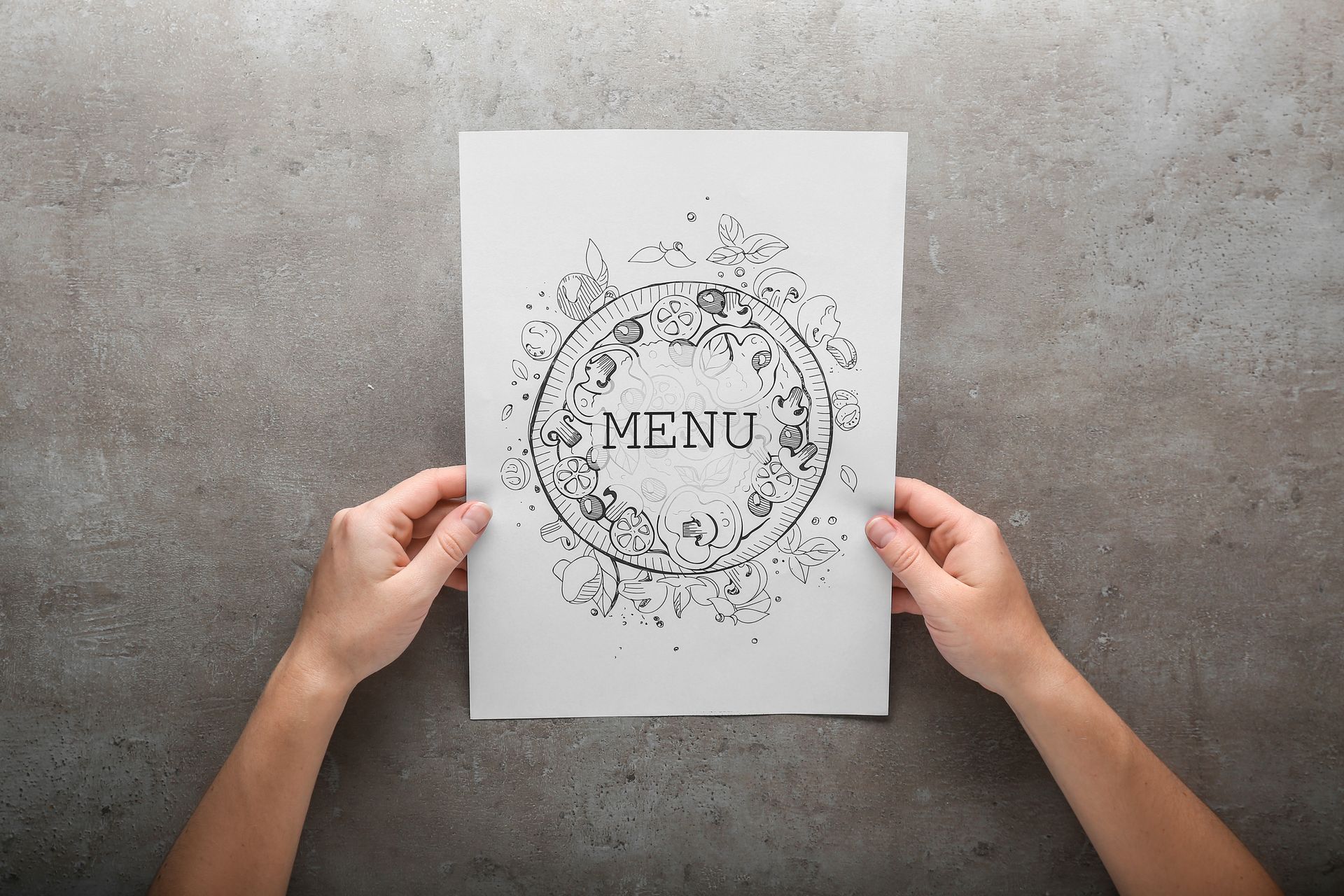 Des mains tiennent un menu sur une surface grise où figure le mot MENU dans un motif floral circulaire.