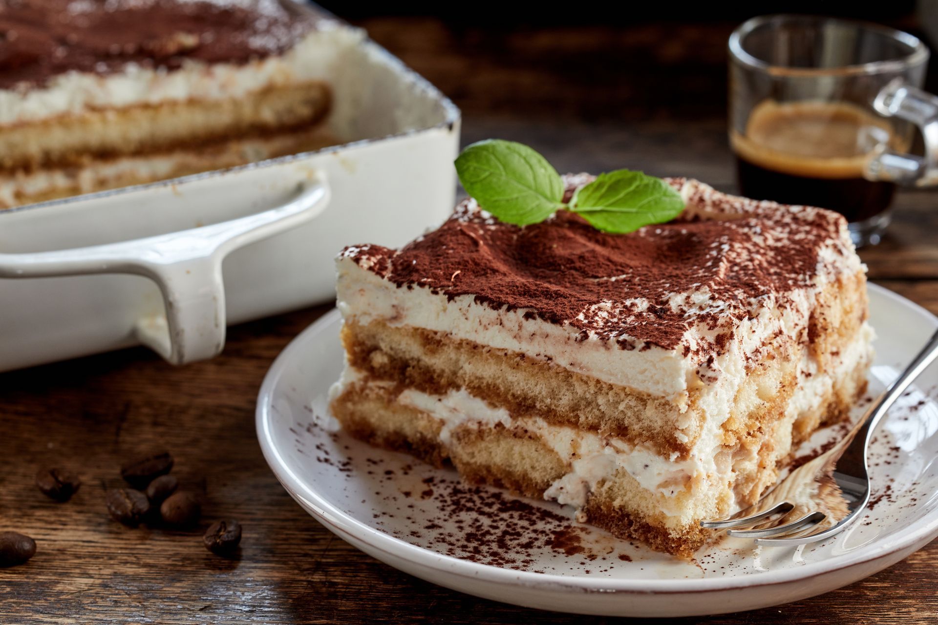 Une part de tiramisu sur une assiette blanche, saupoudrée de cacao et agrémentée d'un brin de menthe ; du café en arrière-plan.