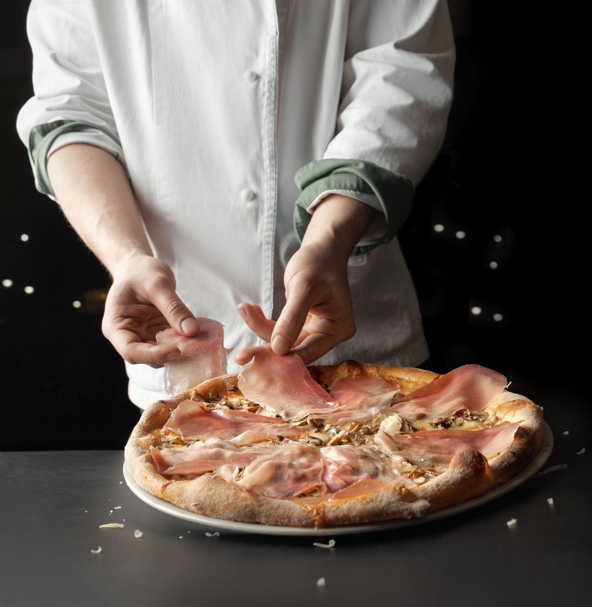 Un chef dépose des tranches de prosciutto sur une pizza, sur fond sombre.