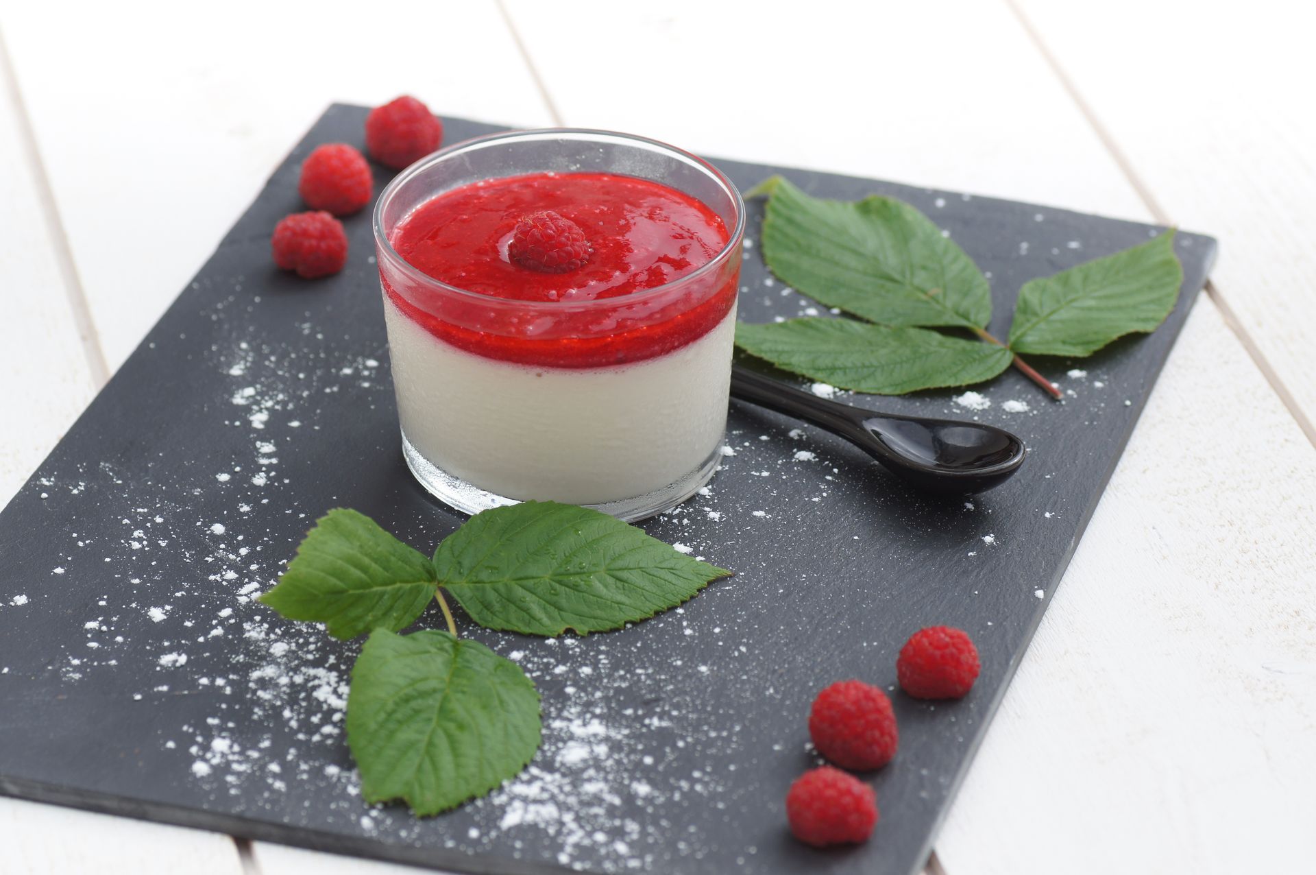 Panna cotta servie dans un verre avec une sauce aux framboises, sur une ardoise noire, avec des framboises et des feuilles vertes.