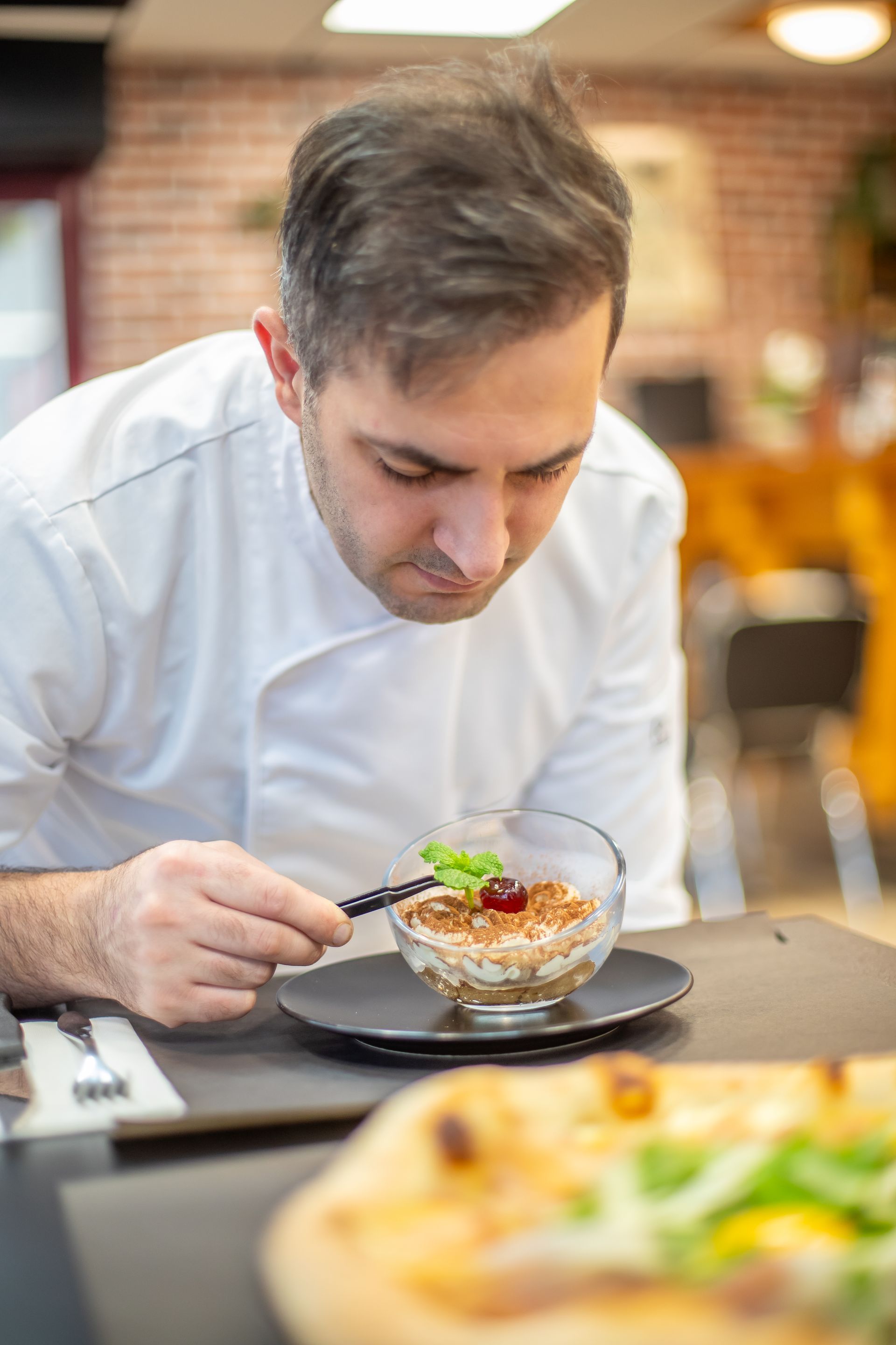 Une part de tiramisu sur une assiette blanche, saupoudrée de cacao et agrémentée d'un brin de menthe ; du café en arrière-plan.