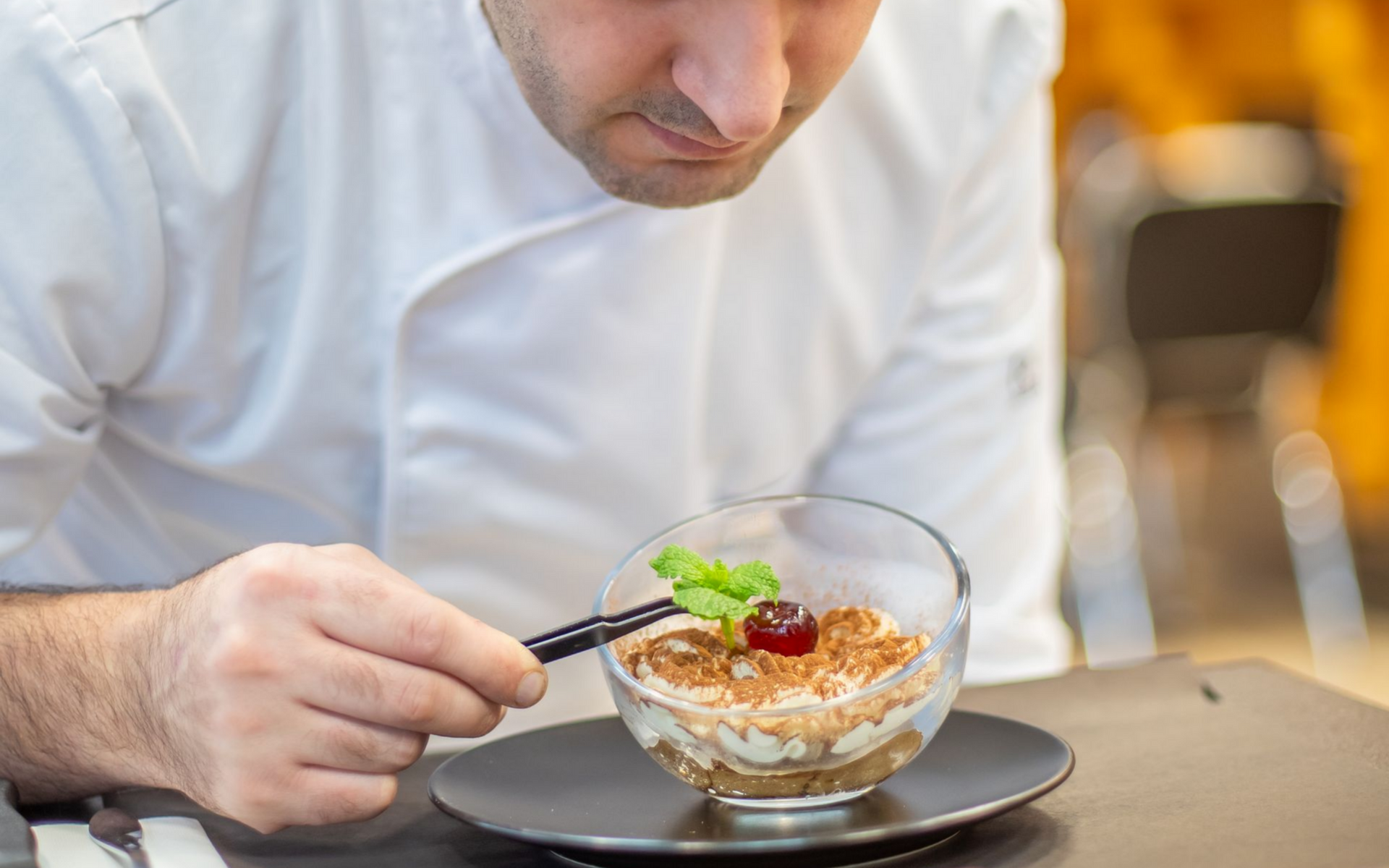 Une part de tiramisu sur une assiette blanche, saupoudrée de cacao et agrémentée d'un brin de menthe ; du café en arrière-plan.
