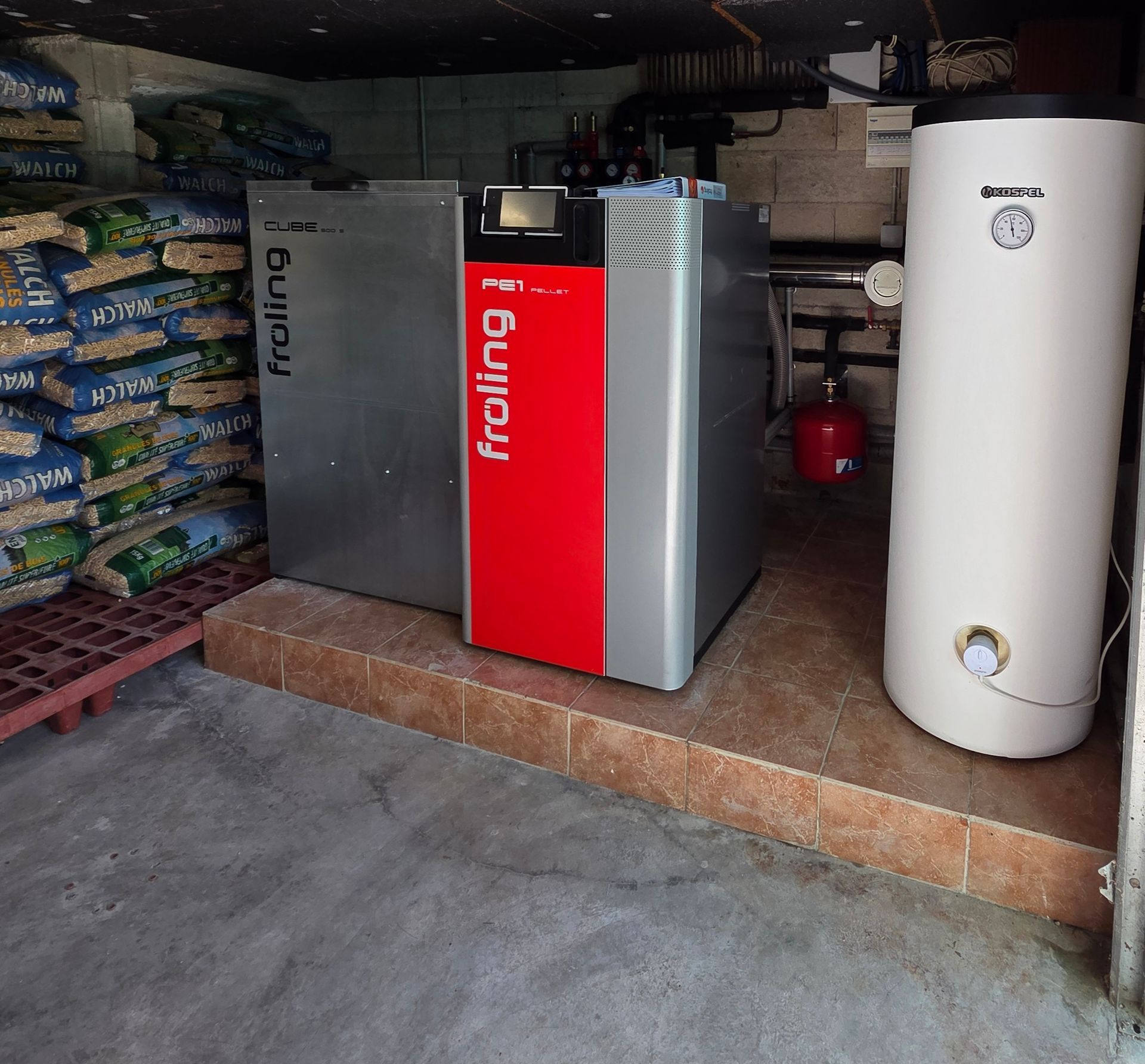 Un système de chauffage avec une chaudière à granulés rouge et grise et un réservoir d'eau blanche dans un environnement de sous-sol.