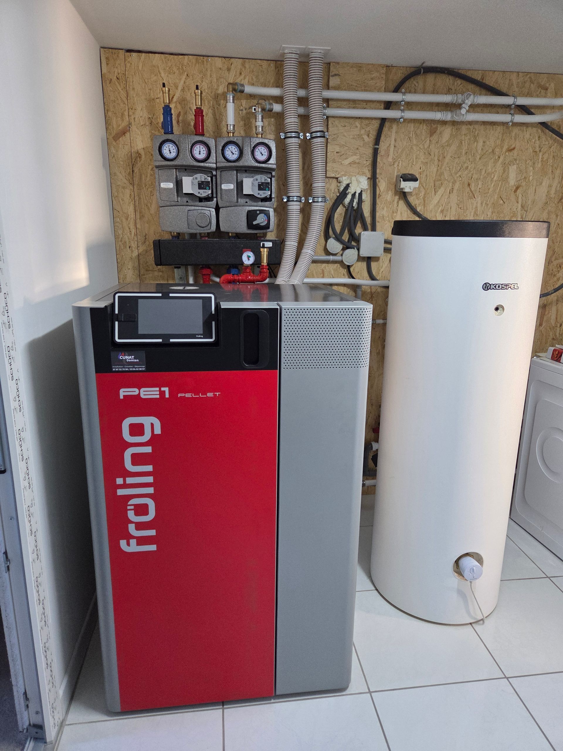 Une chaudière à granulés Fröling rouge et grise avec un réservoir d'eau blanche dans une buanderie.