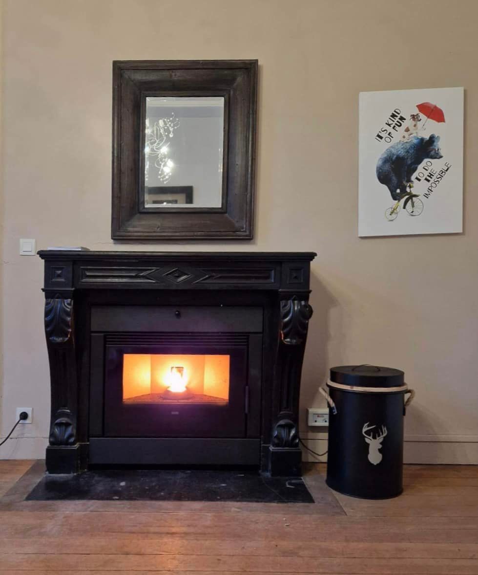 Cheminée noire avec flammes, miroir et œuvres d'art sur un mur beige. Un récipient noir est posé sur le côté.