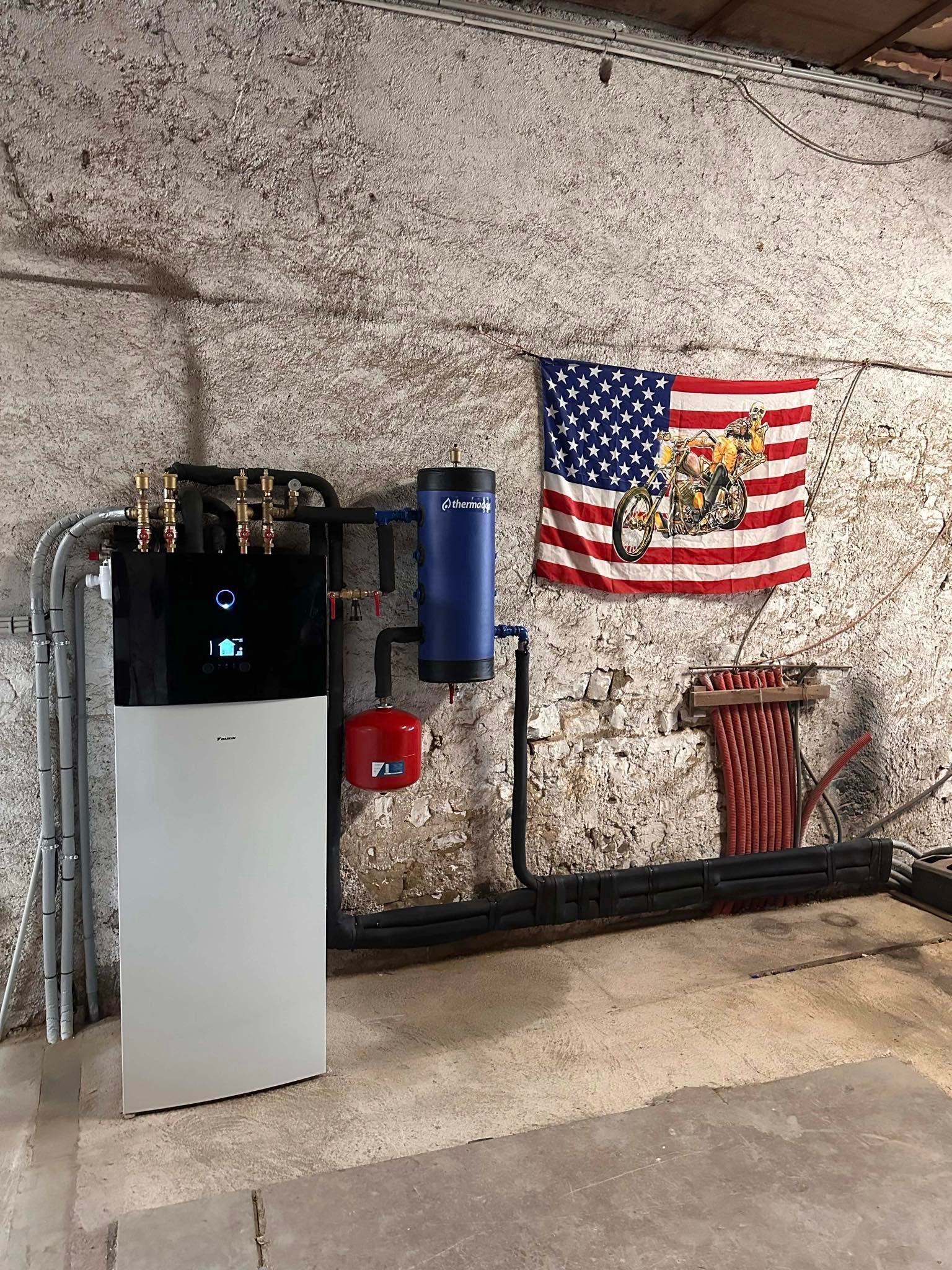 Composants du système de chauffage contre un mur en pierre patiné, comprenant une unité blanche, un réservoir bleu et un composant rouge, avec un drapeau.