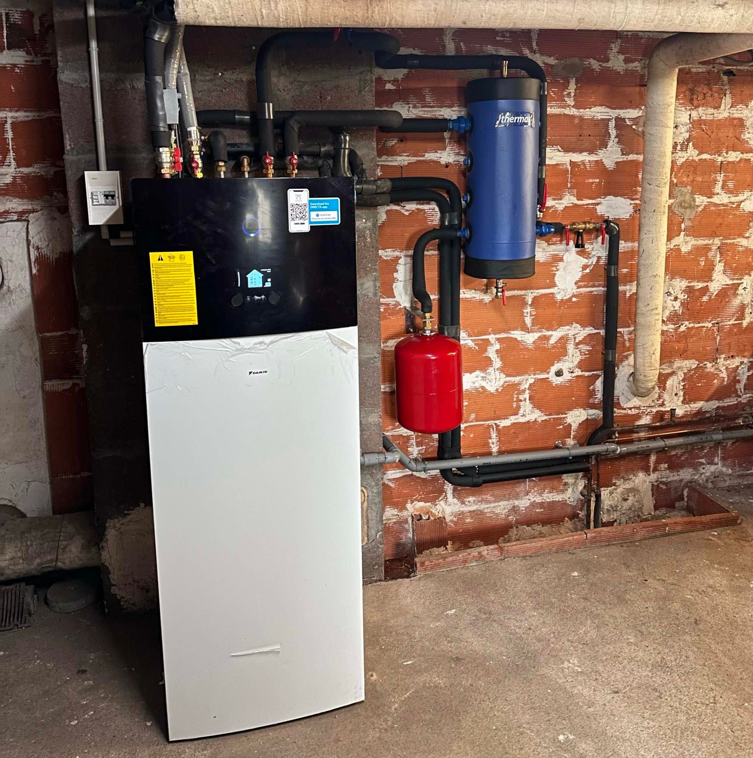 Système de chauffage avec un réservoir noir, un panneau blanc et un cylindre bleu sur un mur de briques.