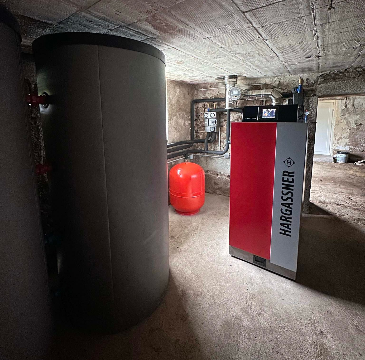 Système de chauffage au sous-sol avec réservoir d'eau, chaudière Hargassner, tuyauterie et vase d'expansion.