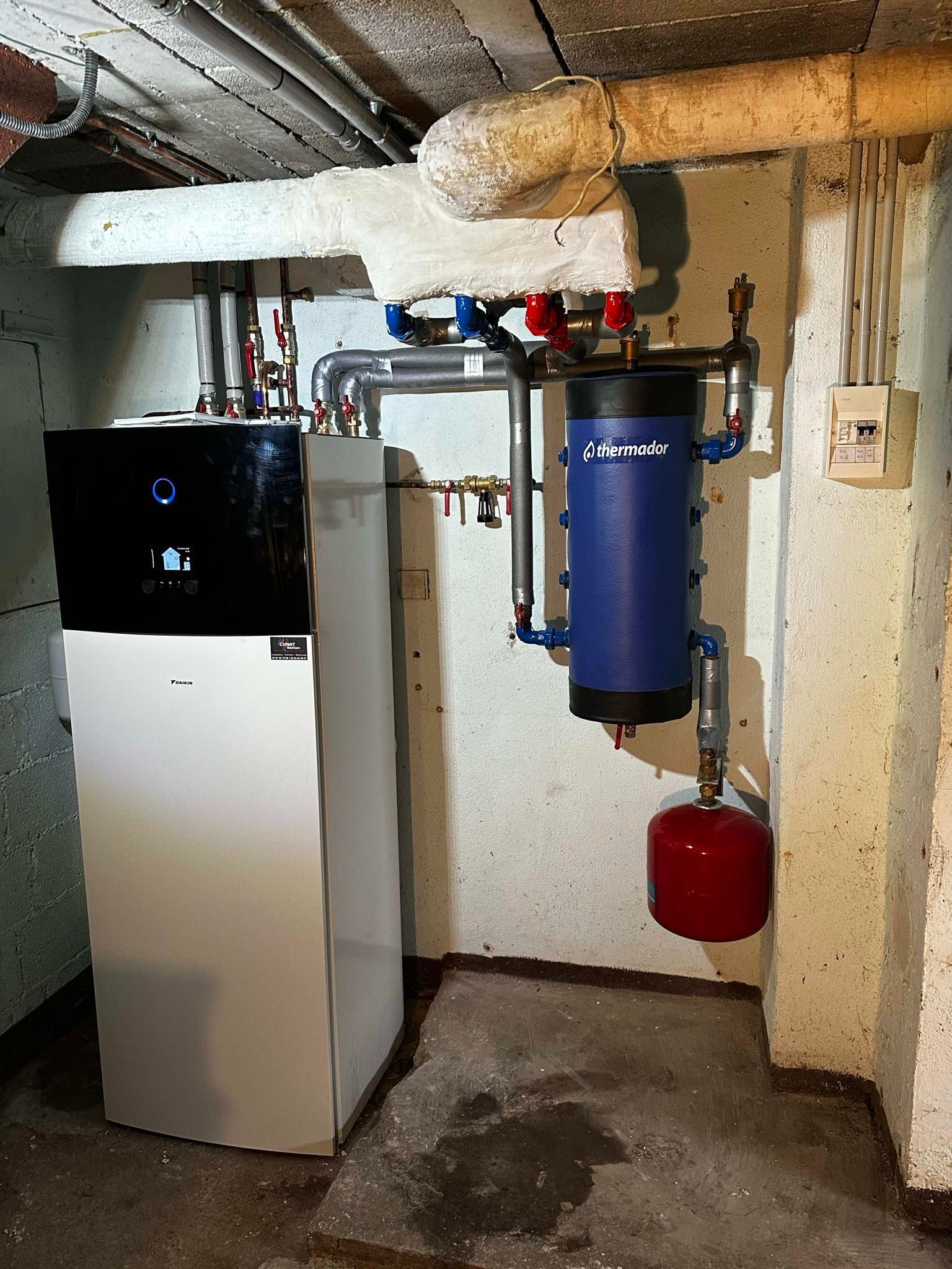 Système de chauffage au sous-sol avec unité blanche, réservoir bleu, vase d'expansion rouge et tuyaux.