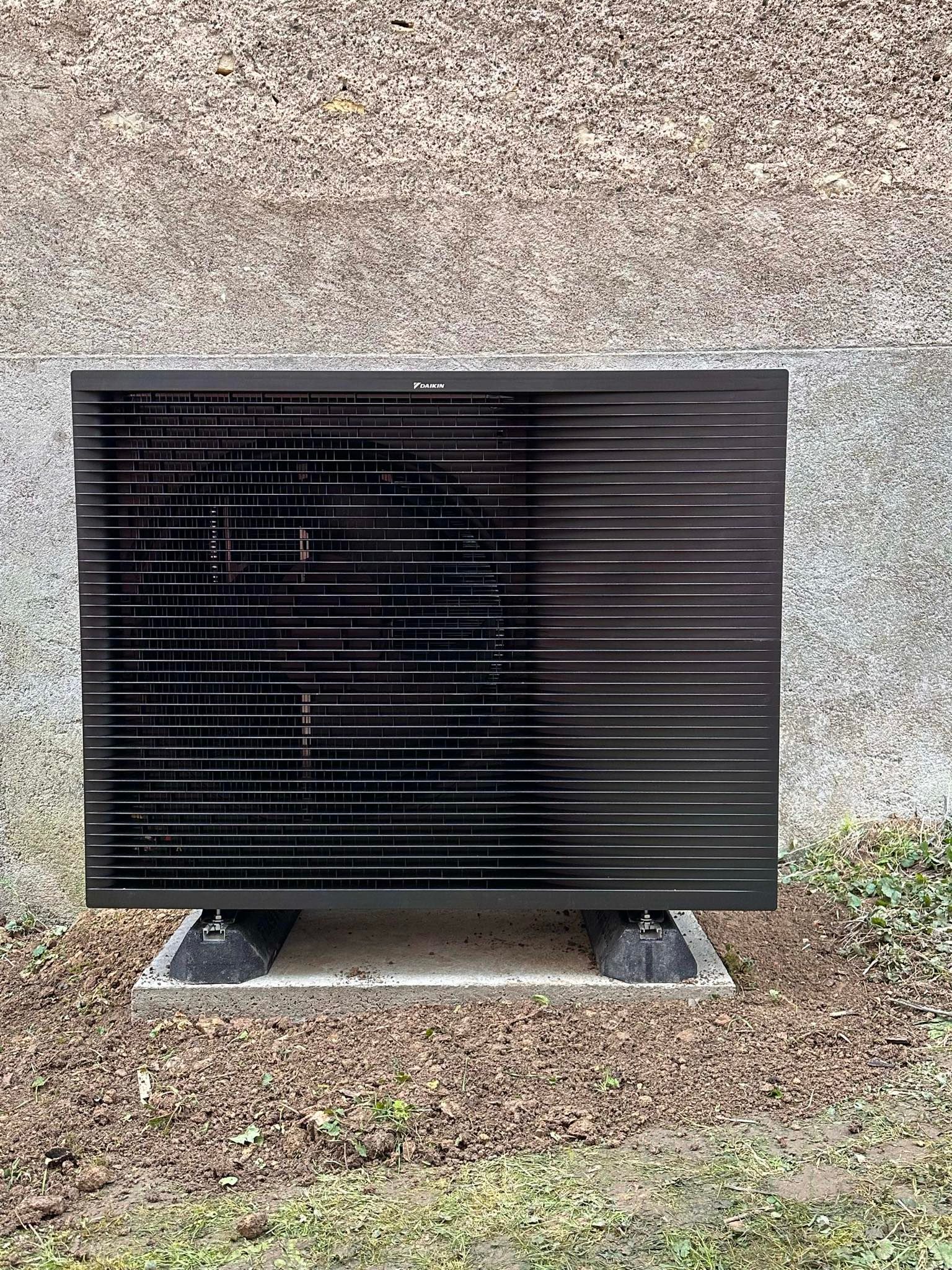 Unité extérieure rectangulaire noire, sur une base en béton contre un mur texturé.