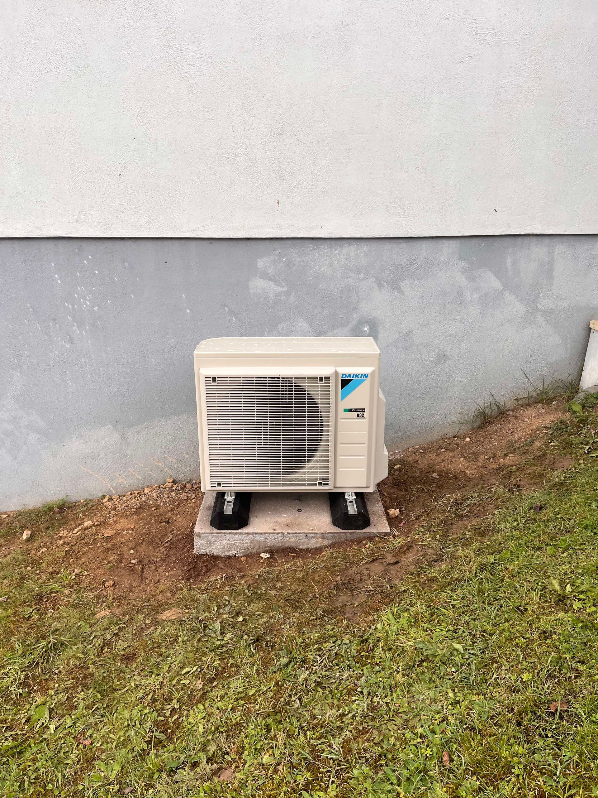 Une unité de climatisation extérieure sur une base en béton, à proximité d'un bâtiment blanc avec de l'herbe devant.