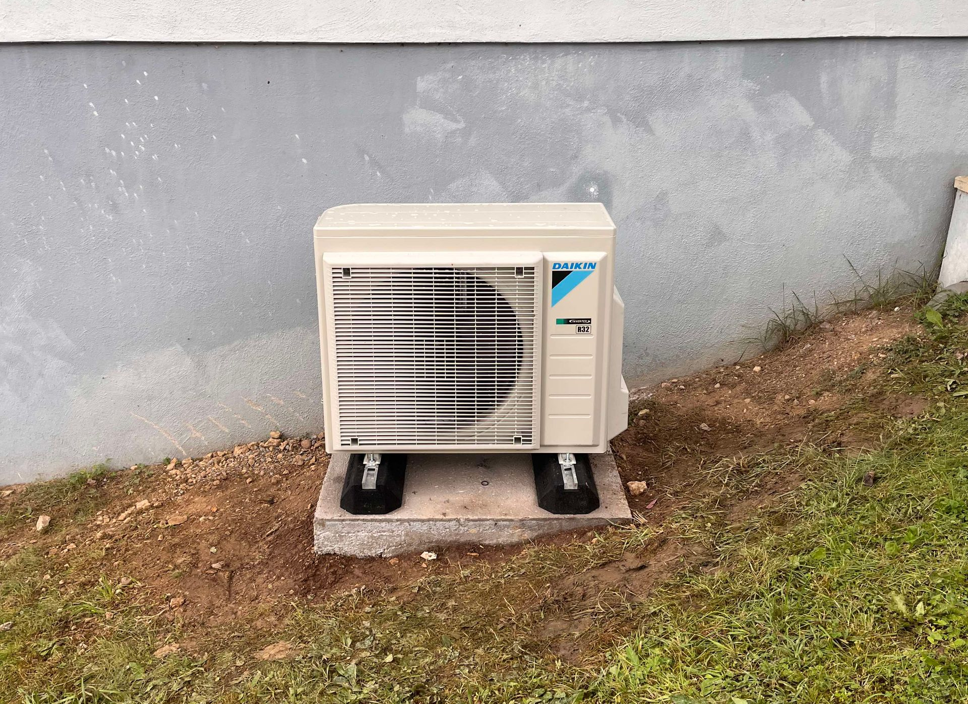 Unité de climatisation Daikin montée sur une dalle de béton près du mur extérieur d'un bâtiment et de l'herbe.