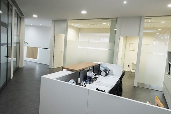 Luminosa zona de recepción de oficinas con escritorios blancos, paredes de cristal e iluminación cenital.