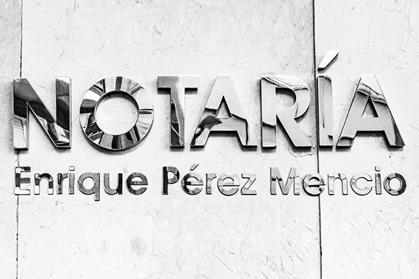 Metal sign reading “NOTARÍA Enrique Pérez Mencía” on a white wall.