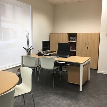 Oficina vacía con escritorio, ordenador, armarios, sillas y una mesa redonda junto a la ventana.