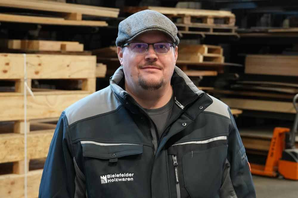 Ein Mann mit Hut und Brille steht vor Holzpaletten.