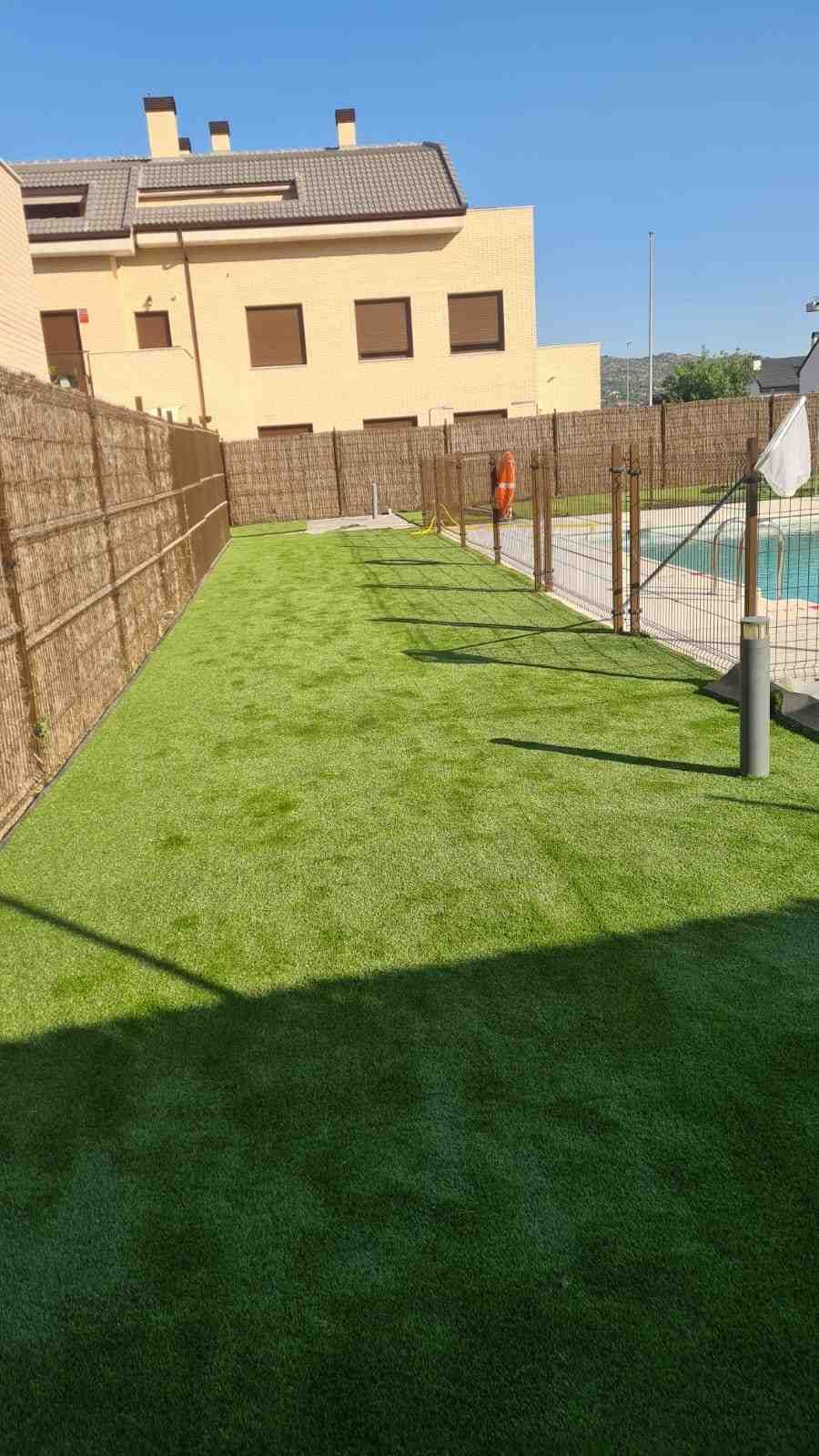 Un césped de un verde brillante junto a una piscina vallada, con un edificio de varias plantas al fondo.