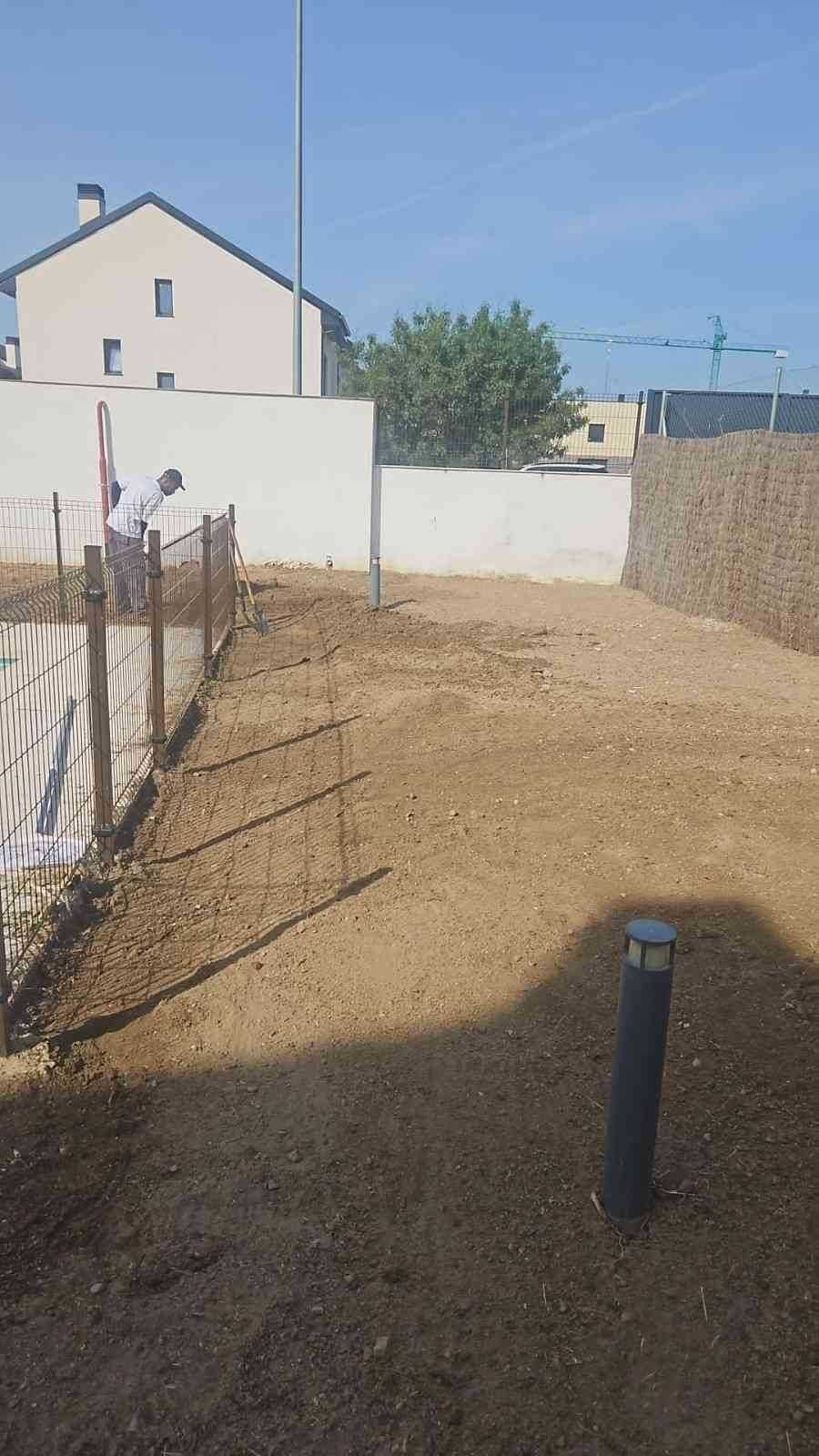Una persona se encuentra de pie en un patio de tierra junto a una cerca de alambre, con una casa y paredes blancas al fondo.