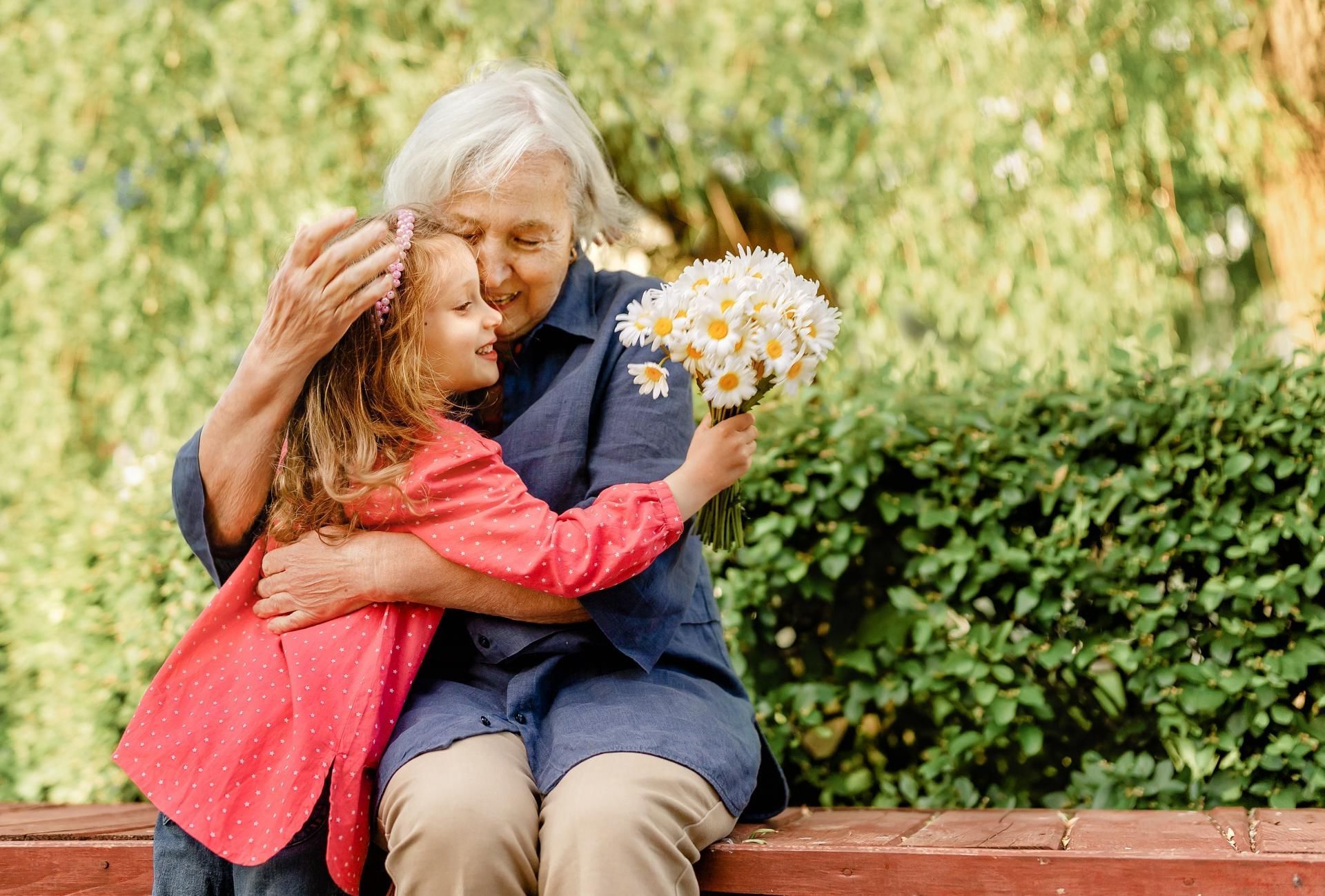 Eine ältere Frau sitzt mit einem kleinen Mädchen auf einer Bank und hält einen Strauß Gänseblümchen.