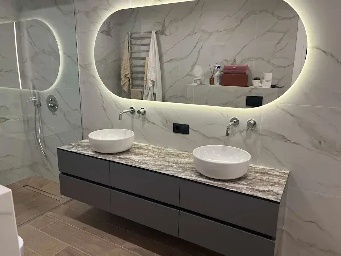 Tocador de baño moderno con dos lavabos sobre encimera, encimeras de mármol y espejos ovalados retroiluminados.