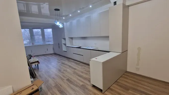 Cocina moderna vacía con armarios blancos, suelos de madera y una luminosa zona de estar diáfana.