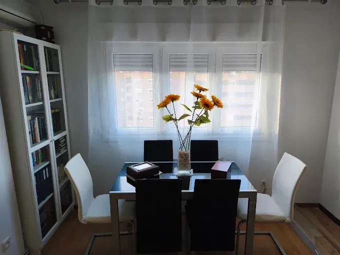 Comedor con mesa negra, sillas blancas, jarrón con flores amarillas y ventanas con cortinas transparentes.