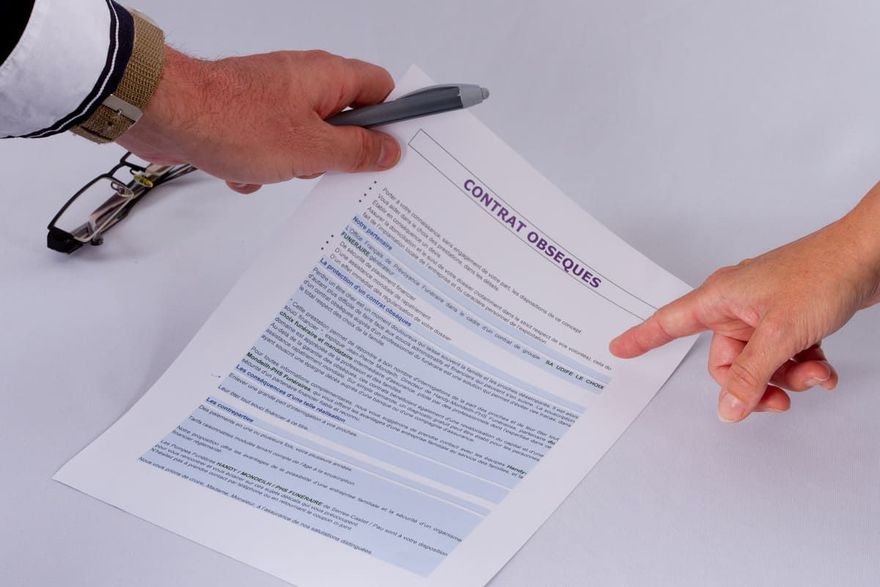 Des mains examinent un contrat ; une main désigne du doigt tandis que l’autre tient un stylo. Des lunettes sont posées à proximité sur une surface blanche.