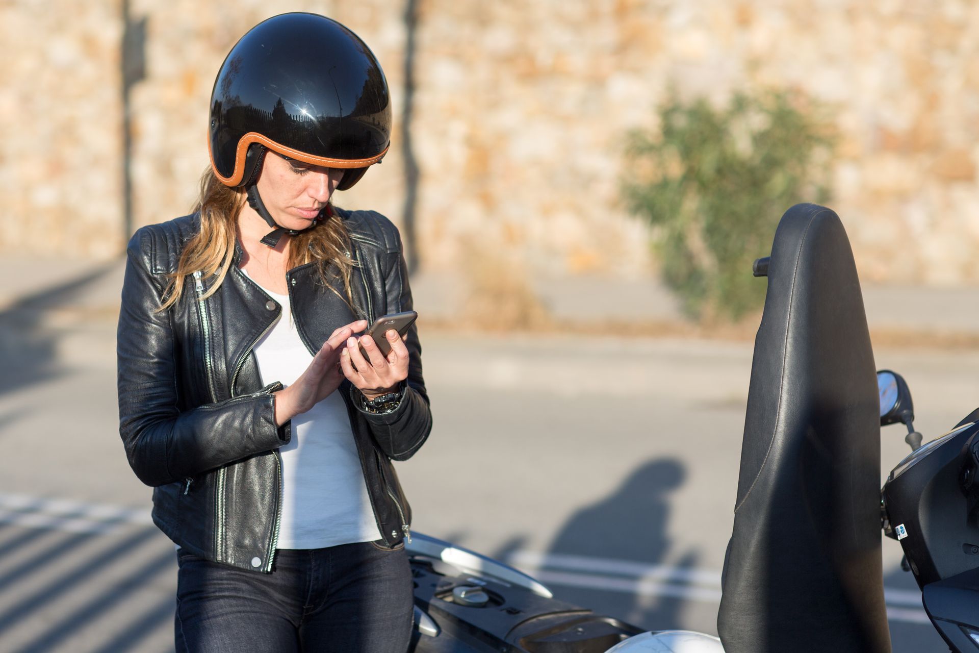 Une femme portant un casque et une veste utilise son téléphone à côté d'un scooter.