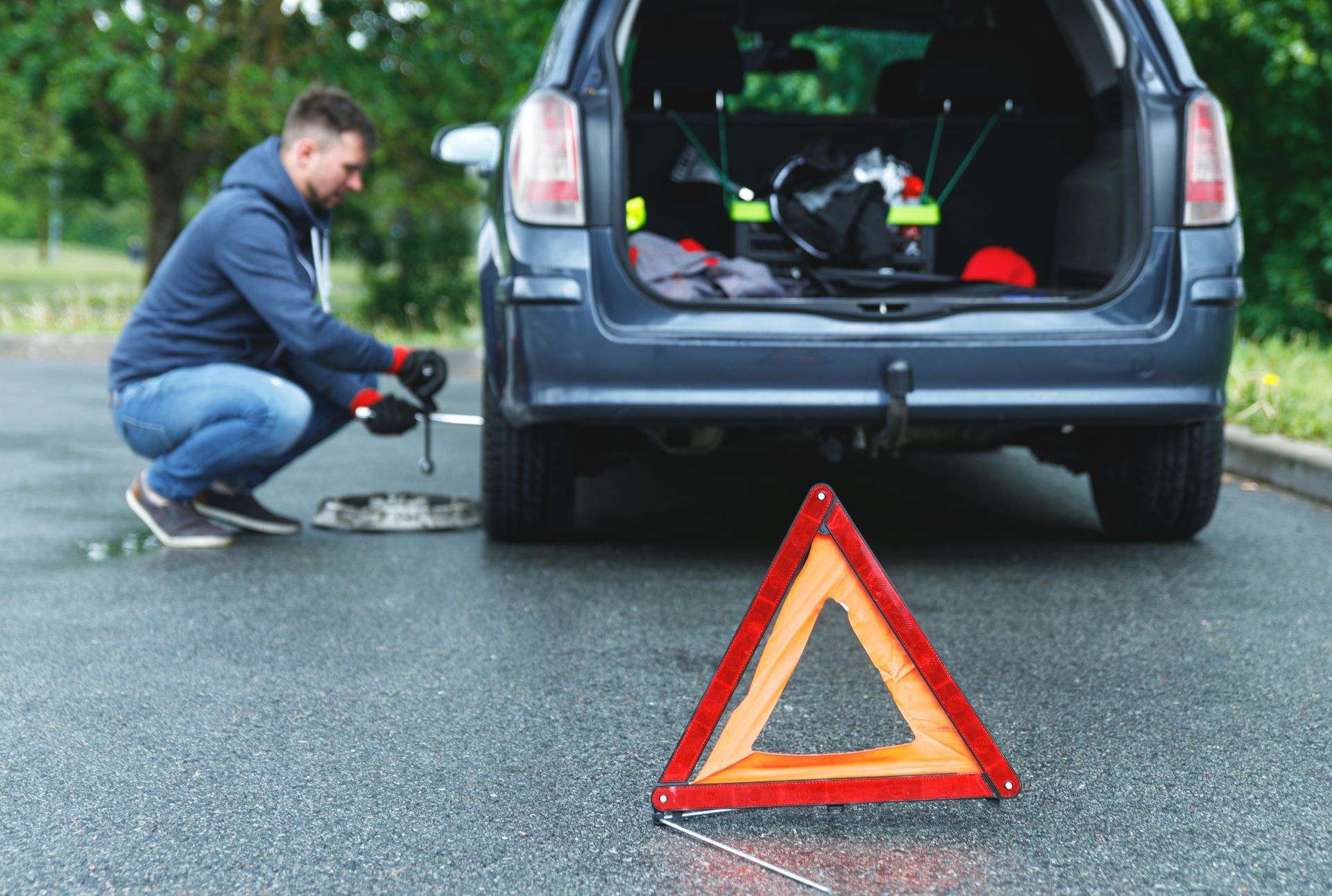 Un homme change un pneu crevé sur une voiture garée sur le bord de la route ; un triangle orange de signalisation est visible devant.