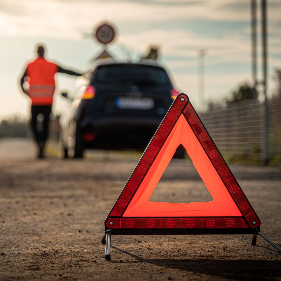Triangle de signalisation orange sur la route. Une personne portant un gilet de sécurité se trouve près d'une voiture.