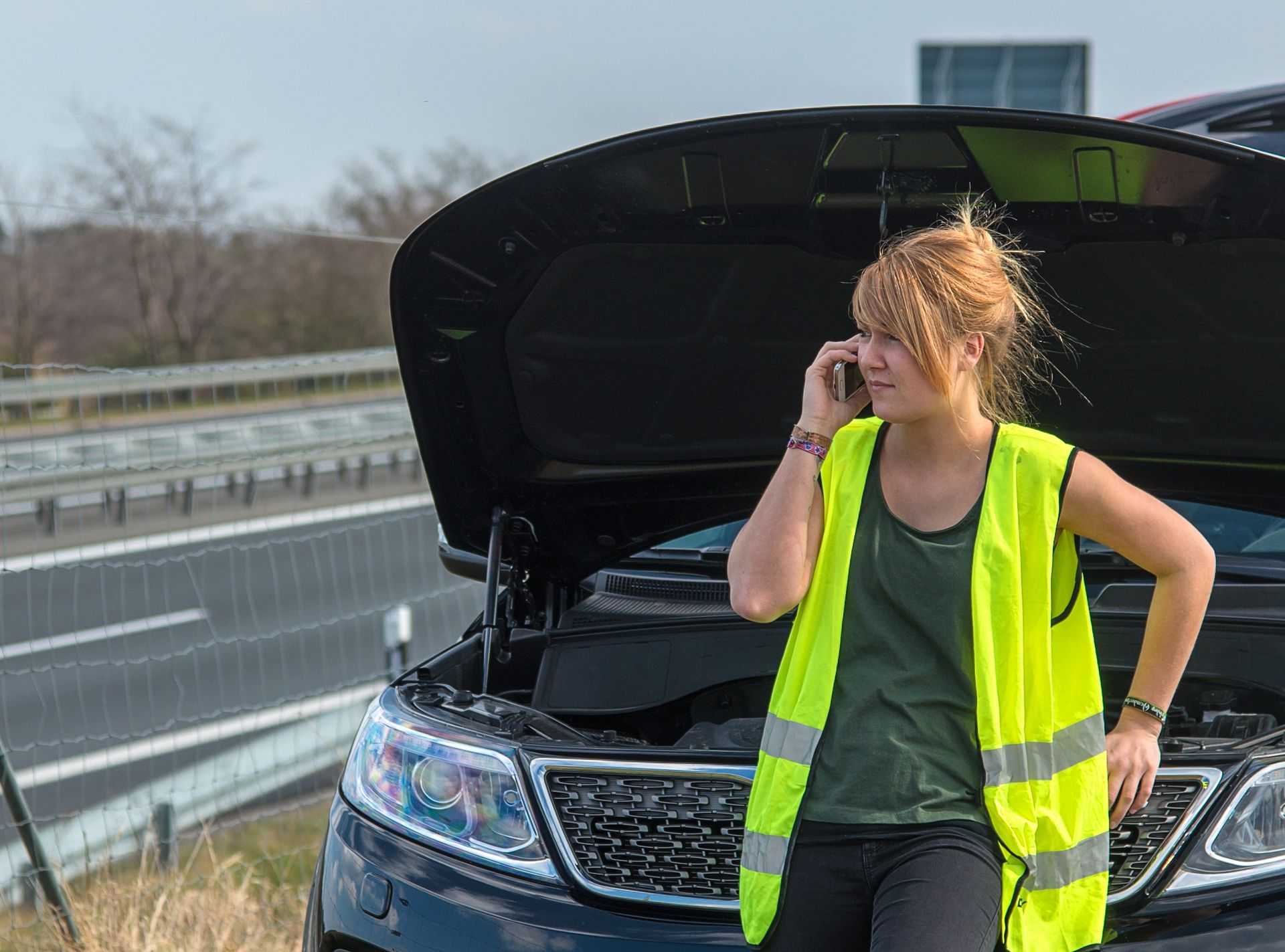 Une femme en gilet jaune parle au téléphone à côté d'une voiture avec le capot ouvert sur le bord d'une autoroute.