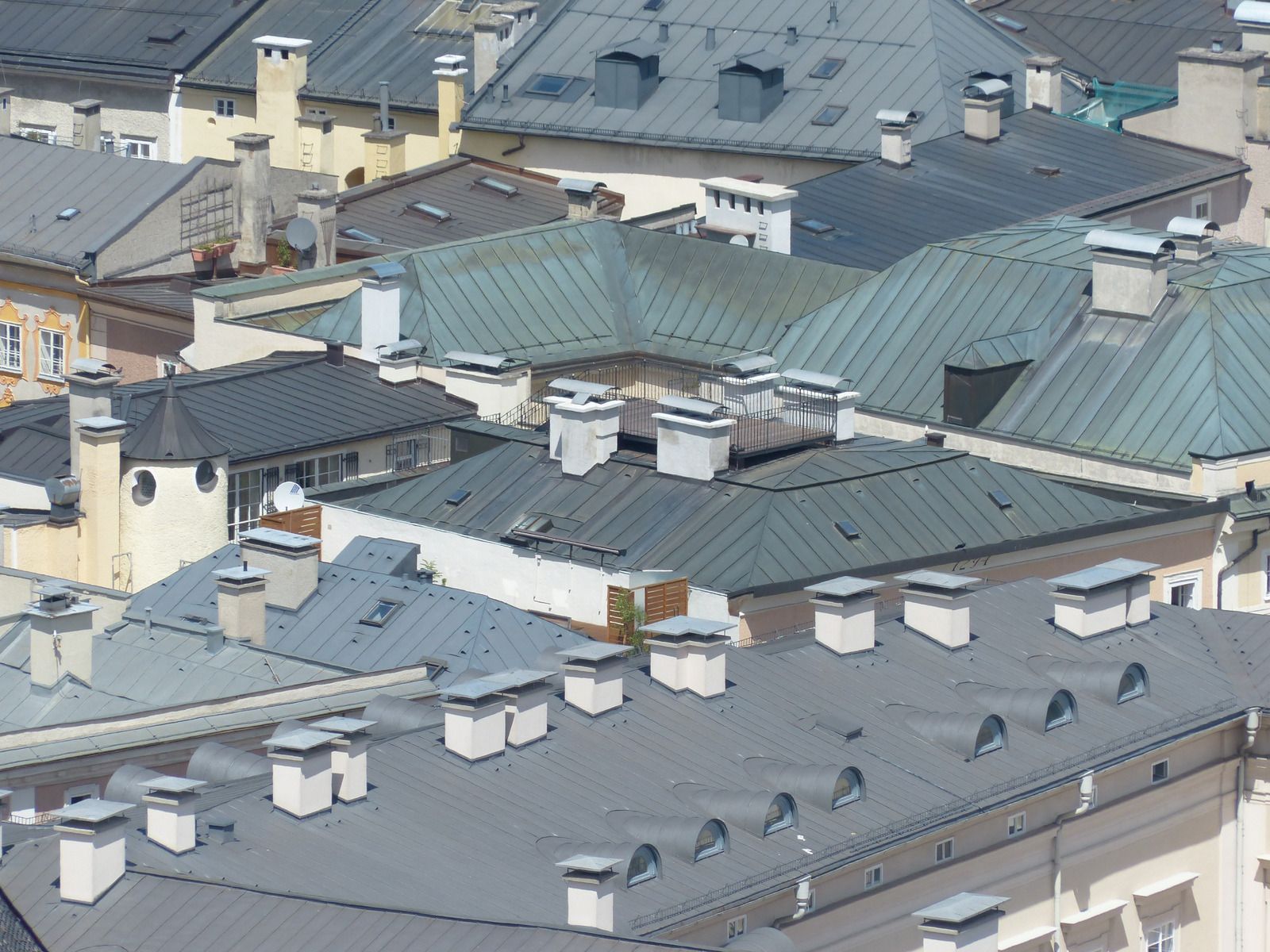 Rooftops with gray and green metal roofs and multiple white chimneys in a dense urban neighborhood