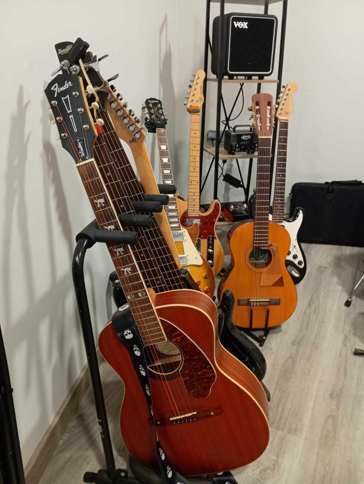 Varias guitarras acústicas y eléctricas en soportes dentro de una habitación, con un pequeño amplificador Vox al fondo
