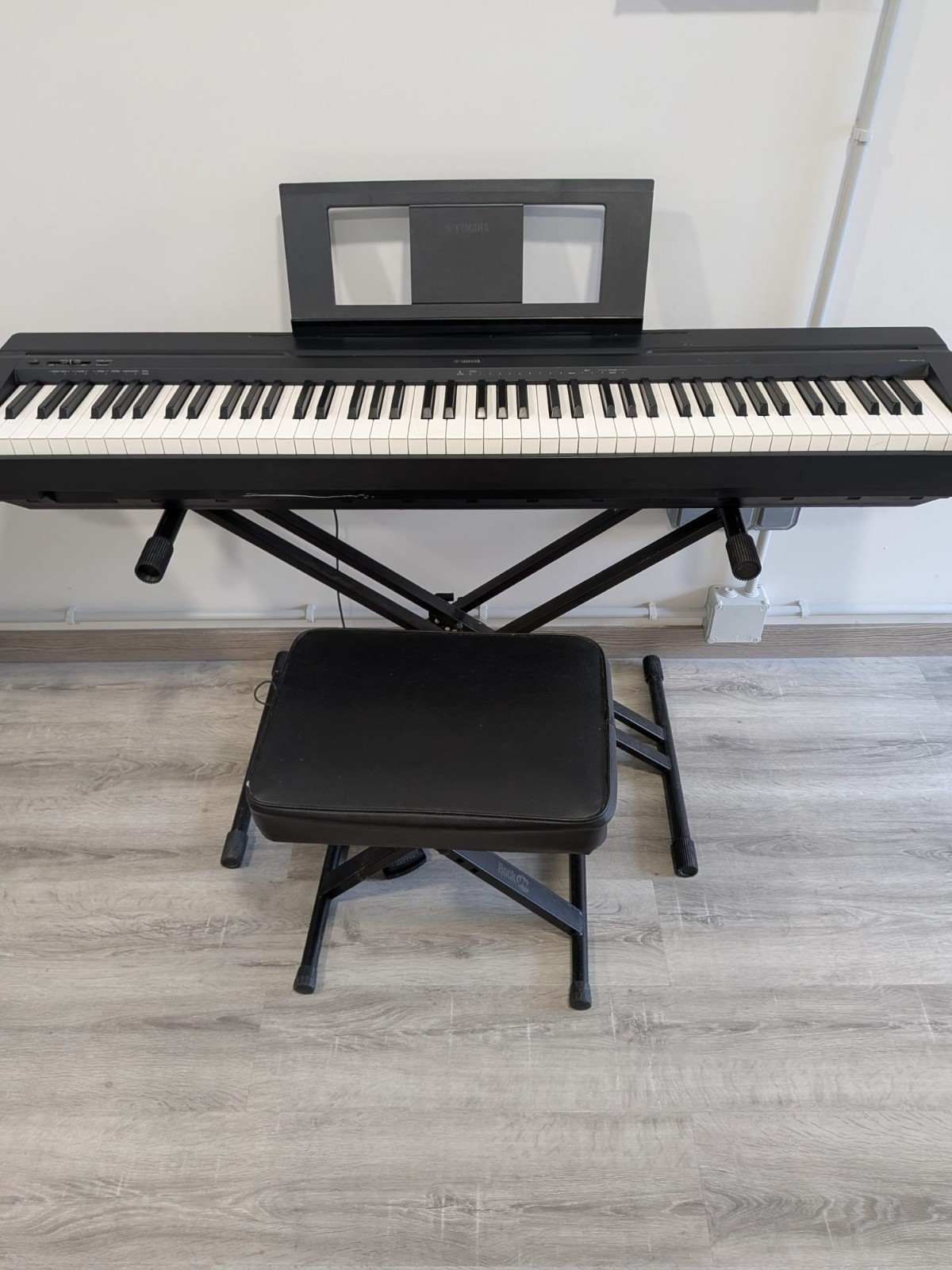 Piano digital negro con banqueta apoyado contra la pared en una habitación luminosa