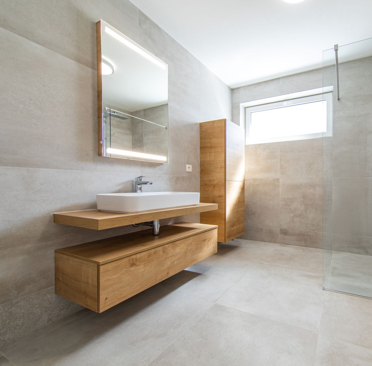 Une salle de bains moderne avec des murs en pierre claire, un meuble lavabo suspendu avec un lavabo, un miroir éclairé et une armoire haute.