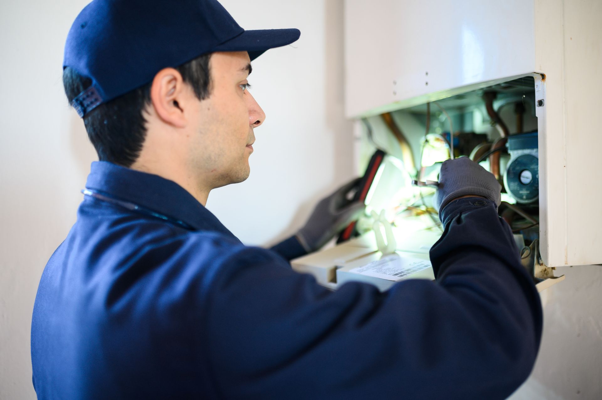 Un technicien en combinaison et casquette inspecte les composants internes d'une chaudière murale à l'aide d'une lampe portative.