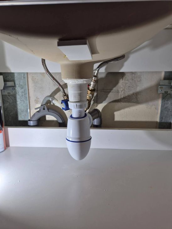 Siphon d'évier en plastique fixé à un lavabo dont les conduits d'alimentation métalliques sont visibles en arrière-plan.