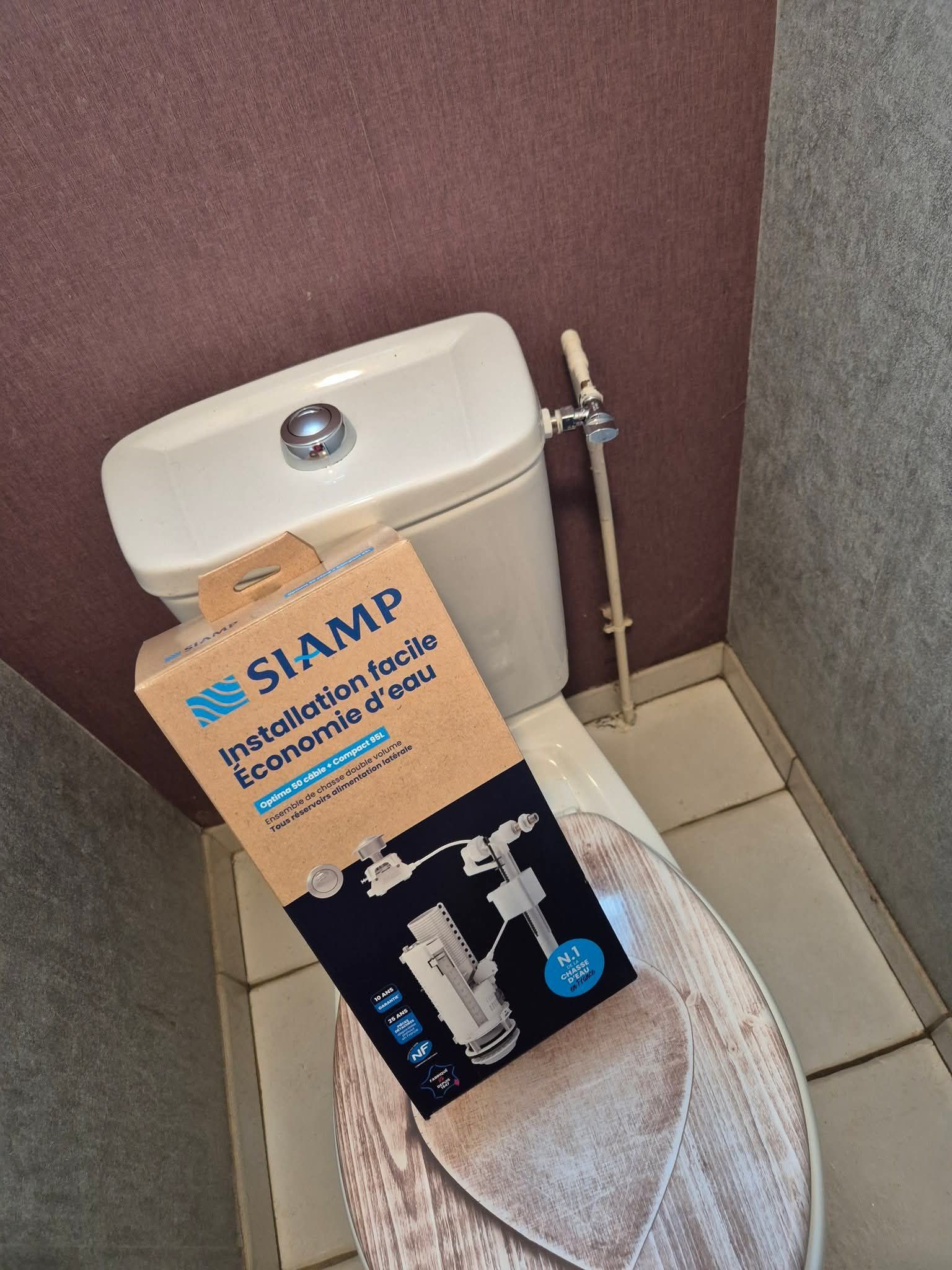 Dans une salle de bains carrelée, une boîte en carton contenant un mécanisme de chasse d'eau SIAMP à économie d'eau est posée sur des toilettes à réservoir.
