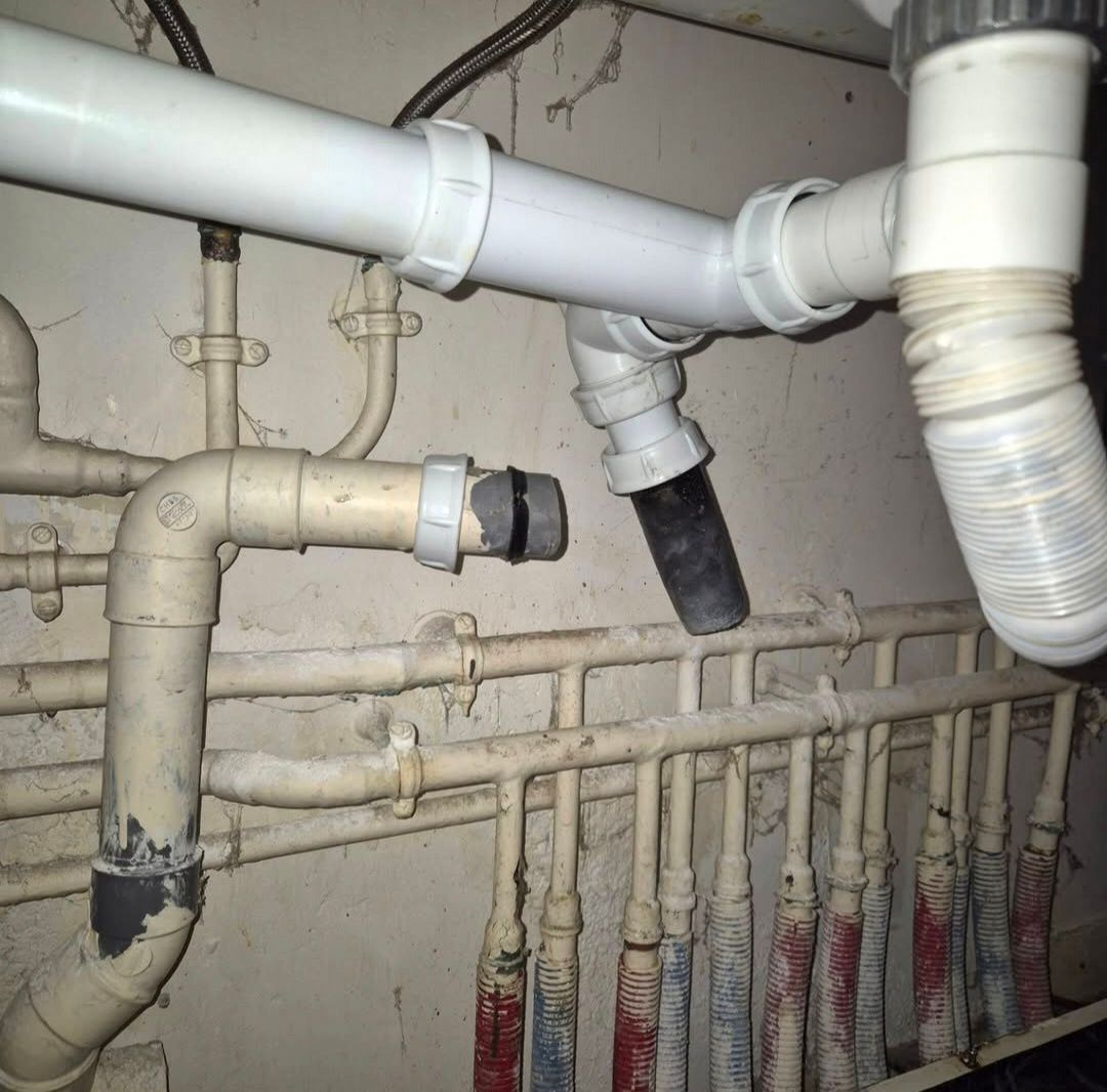 Vue sous un évier montrant des tuyaux d'évacuation en plastique déconnectés, un raccord en caoutchouc et plusieurs conduites d'eau.